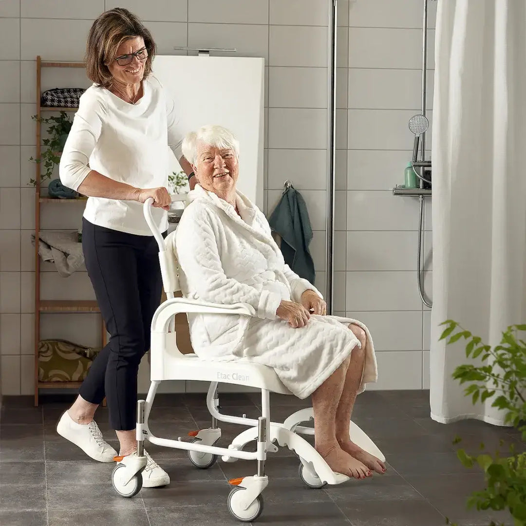 Woman assisting an elderly woman in a bathrobe using a white bath chair in a bathroom.