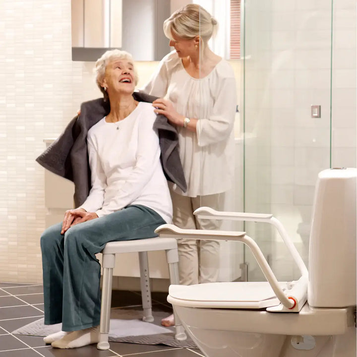 Woman assisting an elderly woman on a shower chair in a bathroom.