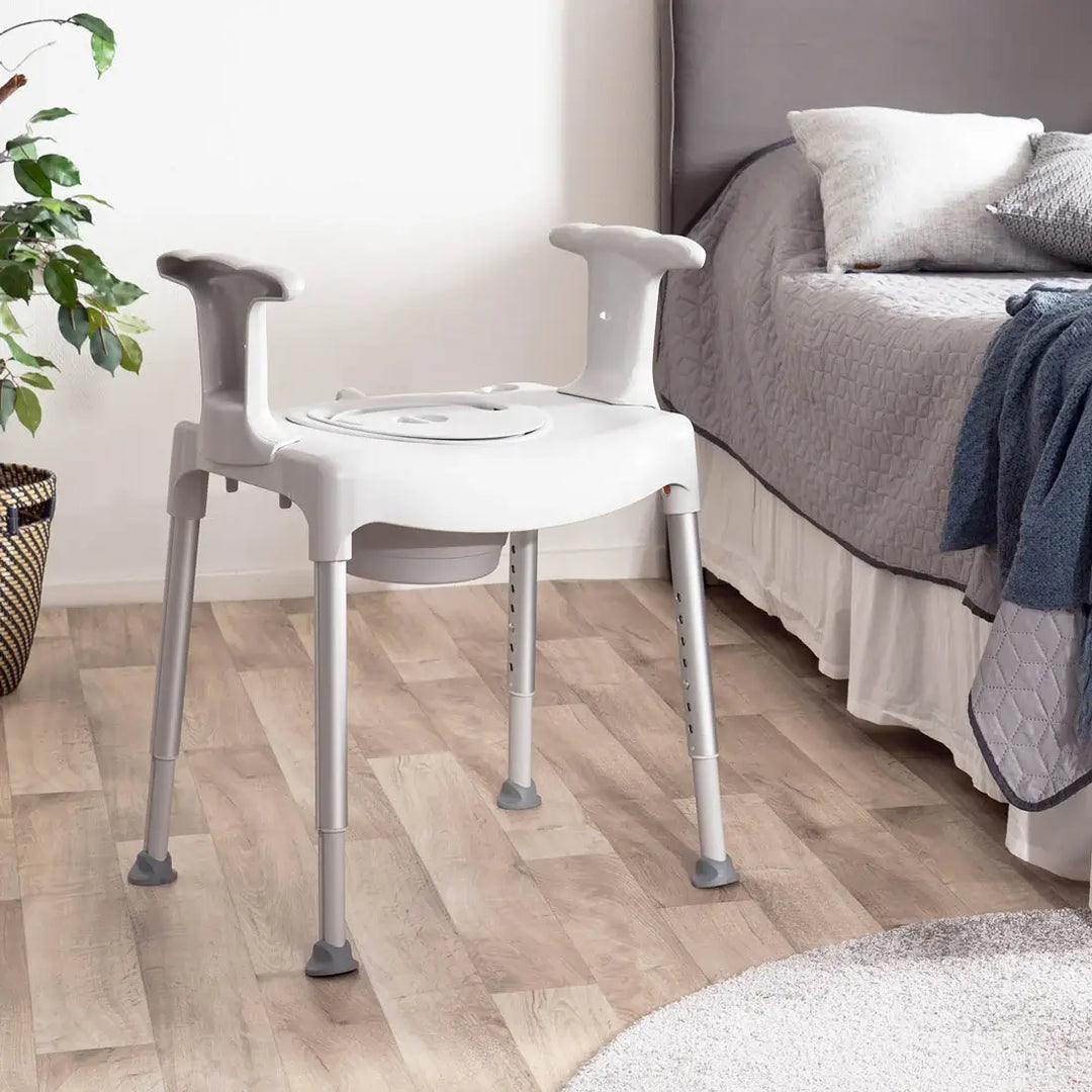 White commode chair on a wooden floor next to a bed with gray bedding.