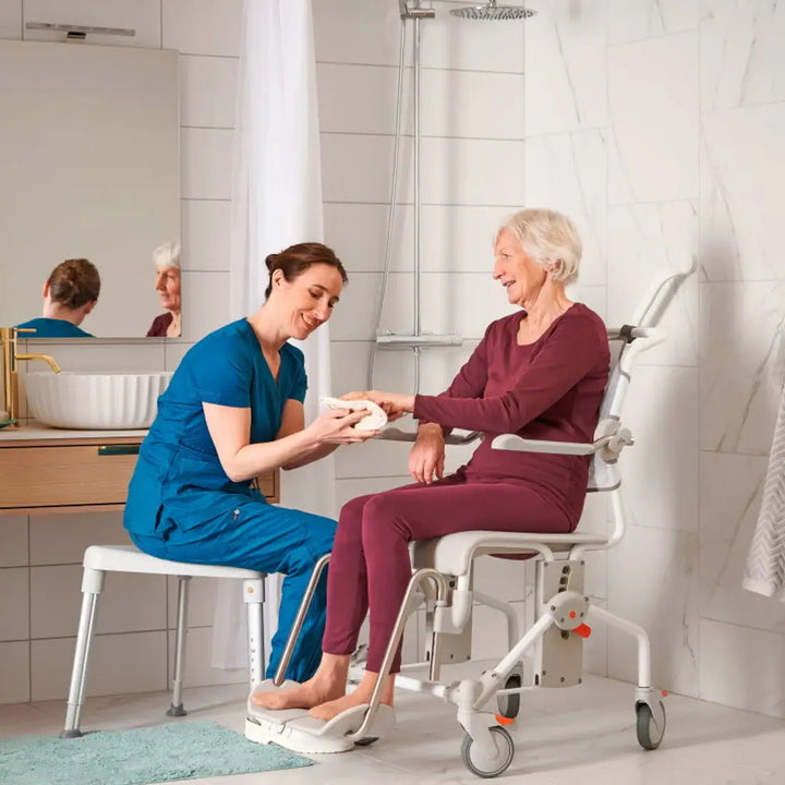 Woman assisting an elderly woman using a shower chair in a bathroom.